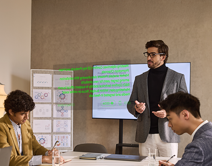 A man wearing INMO GO3 speaks in front of a screen in a meeting room, with green script text floating in front of him and two colleagues seated at a table