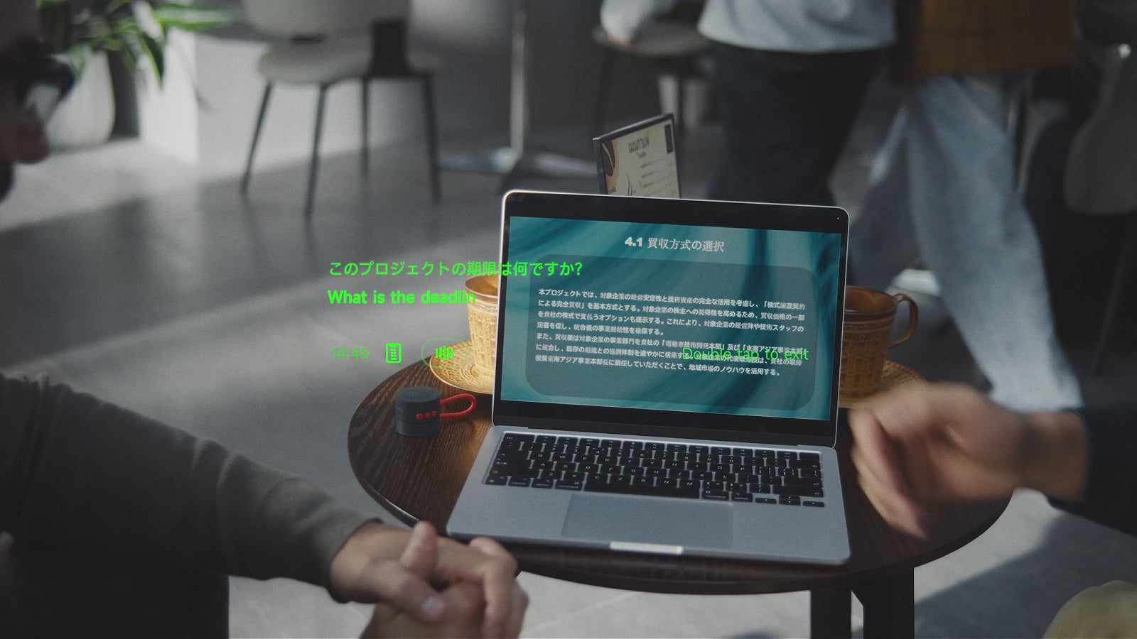 Two people talk at a table with a laptop and INMO Speaker while a Japanese question and its English translation, What is the deadline, appear in green text